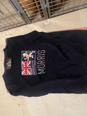 Svart t-shirt från Morris med tryck - Snygg svart t-shirt från Morris med stort tryck på bröstet som visar en brittisk flagga, ett lejon och texten 'Morris'. T-shirten har rund hals och korta ärmar. Perfekt för dig som gillar klassisk stil med en twist.