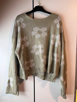 Grön stickad tröja med blommor - Snygg grön stickad tröja med vita blommor i ett oversized snitt. Tröjan har rund halsringning och långa ärmar. Perfekt för dig som gillar en avslappnad och trendig stil.