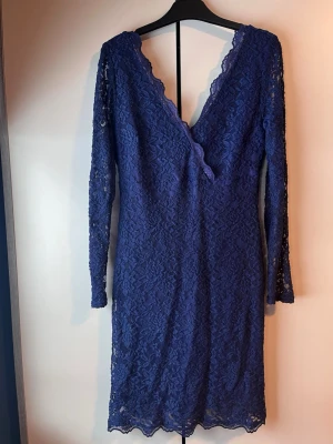 Blå spetsklänning med v-ringning - Säljer en blå spetsklänning med lång ärm och djup v-ringning både fram och bak. Klänningen har en figurnära passform och är täckt av ett vackert spetsmönster. Perfekt för dig som vill ha en elegant look med lite extra detaljer. Supersexig och sitter sååå snyggt på. Använd endast en gång. Lite för kort för mig.