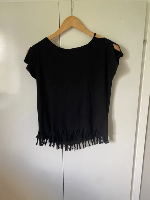 Svart topp med fransar - Svart kortärmad topp med rund halsringning och fransar längs nederkanten. Toppen har en lös passform och är tillverkad i mjuk bomull, perfekt för en avslappnad och trendig stil. Skulle säga en liten L, 