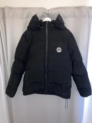 Svart pufferjacka från Trapstar - Säljer en svart pufferjacka från Trapstar med huva och dragkedja framtill. Jackan har en rund logga på bröstet och Trapstar-broderi i stor stil på ryggen. Fylld för extra värme och har två fickor med dragkedja. Perfekt för kalla dagar och streetwear-stil.