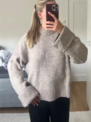Beige oversized stickad tröja - Beige stickad tröja med oversized passform och breda ribbade muddar vid ärmslut och nederkant. Tröjan har rund halsringning och är gjord i ett mjukt, tjockt material som ger en avslappnad look.