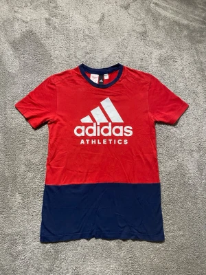 Röd och marinblå Adidas t-shirt - Snygg röd t-shirt från Adidas med stor vit logga och texten 'ATHLETICS' på bröstet. T-shirten har en marinblå nederdel och matchande marinblå krage. Klassisk passform och korta ärmar, perfekt för en sportig look.