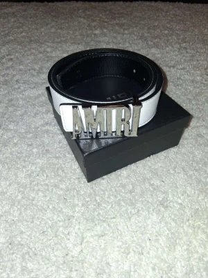 Vitt Amiri bälte med silverdetalj - Säljer ett stilrent vitt bälte från Amiri med stor silverfärgad logotyp som spänne. Bältet har en klassisk form och är gjort i skinn med svart insida. Perfekt accessoar för att lyfta din outfit.