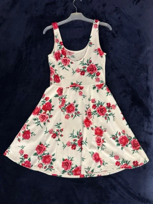 Blommig kortklänning med rosor - Söt vit kortklänning med röda rosor och gröna blad. Klänningen är ärmlös med rund halsringning och har en lätt utställd kjol. Perfekt för varma dagar och somriga tillfällen.