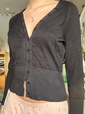 Svart ribbad kofta med v-ringning - Svart kofta med djup v-ringning och knappar framtill. Modellen är figurnära med ribbade partier vid nederkant och ärmslut. Långärmad och tunn i materialet, perfekt för lager-på-lager. Enkel och stilren design som passar till det mesta. Säljer pga för liten🌟osäker på storlek men gissar xs!