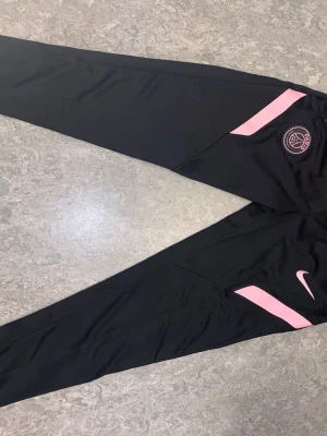 Svarta PSG träningsbyxor Nike - Snygga svarta träningsbyxor från Nike med PSG-logga och rosa detaljer. Byxorna har smal passform, elastisk midja och är perfekta för fotbollsträning eller chill. Materialet är troligtvis polyester för maximal komfort och rörelsefrihet.