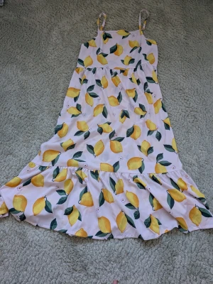 Somrig klänning med citronmönster - Ljus klänning med smala axelband och volang nertill, täckt av ett färgglatt citronmönster. Perfekt för varma dagar och har en avslappnad passform.