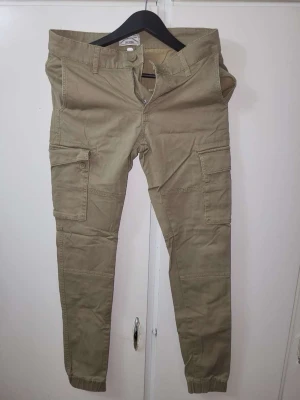 Beige cargobyxor med fickor - Snygga beige cargobyxor med flera praktiska fickor på sidorna och resår vid bensluten. Byxorna har en avslappnad passform och stängs med dragkedja och knapp. Perfekta för en casual och trendig stil.