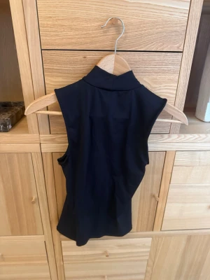 Svart ärmlös polotopp - Snygg svart ärmlös polotopp med tight passform. Perfekt att styla med jeans eller kjol för en clean look. Toppens material känns mjukt och stretchigt, vilket gör den bekväm att bära hela dagen.