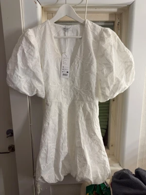 Vit kort klänning med puffärm - Supersöt vit kort klänning från bubbleroom med stora puffärmar, v-ringning och knytband i midjan. Klänningen har ett subtilt blommigt mönster i tyget och volangkant nedtill. Perfekt för studenten! Endast testad men lyckades såklart få en liten sminkfläck som knappt syns men och går säkert bort i tvätten! Slutsåld i denna st på hemsidan