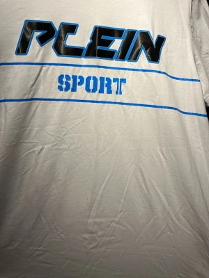 Vit Plein Sport t-shirt med tryck - Snygg vit t-shirt från Plein Sport med svart och blått tryck framtill. T-shirten har korta ärmar och är gjord i mjuk bomull. Detaljer med blå linjer och sportig text ger en cool vibe. Liten etikett med tiger och flaggfärger på sidan.