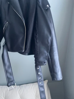 Svart skinnjacka med bälte NA-KD - Snygg svart skinnjacka från NA-KD med klassisk bikerstil, silvriga dragkedjor och ett brett bälte i midjan. Jackan har axelklaffar och en croppad passform som ger en cool vibe. Perfekt för att lyfta vilken outfit som helst. Den har absolut lite skavanker men inget man tänker på tex lite av skinne har flagnat bort nypris 900kr pris kan diskuteras vid snabb och bra affär 