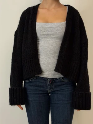 Svart stickad kofta med breda muddar - Mysig svart stickad kofta med öppen front och breda ribbade muddar vid ärmsluten. Koftan har en avslappnad passform och är något croppad, vilket ger en trendig look. Perfekt att slänga över en topp för extra värme.