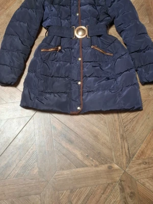 Mörkblå jacka från Attentif - Snygg mörkblå acka från Attentif med bälte i midjan och guldfärgade detaljer. Jackan har två dragkedjeförsedda fickor fram och bruna kantband längs knäppningen. 