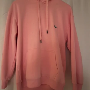 Rosa hoodie från Lacoste - Snygg rosa hoodie från Lacoste med klassisk krokodillogga på bröstet. Hoodien har huva med dragsko, magficka och ribbade muddar vid ärmslut och nederkant. Tillverkad i mjukt bomullsmaterial för en skön känsla.