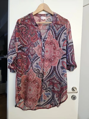 Mönstrad blus från Chica London  - Färgstark blus från Chica London med paisleyinspirerat mönster i rött, blått och vitt. Blusen har v-ringning med knappar framtill och trekvartsärm. Luftigt tunt material som känns skönt mot huden och ger en avslappnad look.