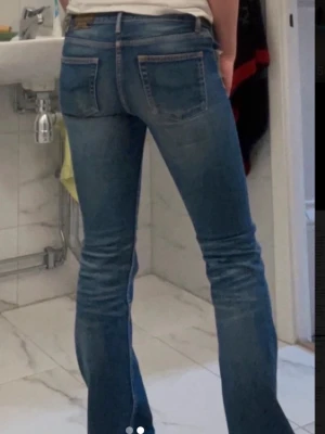 Blå bootcut jeans med slitningar - Säljer ett par blå bootcut jeans med snygga slitningar och tvättade detaljer. Jeansen har klassiska fem fickor och normal midja. Perfekta för dig som gillar en retro och avslappnad stil. Materialet är denim i bomull.