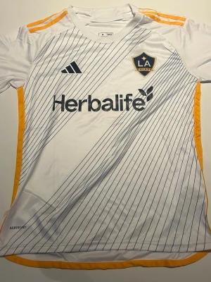 LA Galaxy matchtröja Adidas vit/gul - LA Galaxy matchtröja från Adidas i vitt med tunna diagonala blå linjer, gula detaljer på axlar och sidor samt klubbmärke och sponsortryck på bröstet. Tillverkad i lätt och ventilerande Aeroready-material, perfekt för fotbollsträning eller match.