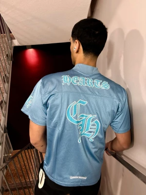Chrome Hearts Jersey Set - Blue - Blå meshtröja från Chrome Hearts med tryck i vitt på bröstet och ryggen. Tröjan har v-ringning, korta ärmar och luftig meshstruktur. Perfekt för en avslappnad streetwear-look.