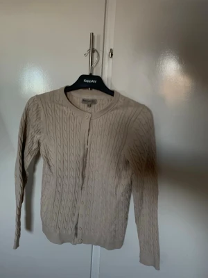 Beige kabelstickad  - Snygg beige kabelstickad kofta från KappAhl med rund halsringning och knappar framtill. Koftan har långa ärmar och ribbade muddar vid ärmslut och nederkant. Perfekt att slänga över en t-shirt eller linne för en chill look.