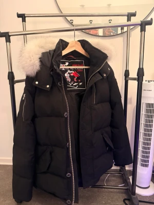  Moose Knuckles vinter jacka - Säljer en svart dunjacka från Moose Knuckles med stor fluffig päls på huvan. Jackan har dragkedja, knappar, fickor och en logotyp på ärmen. Perfekt för kalla vinterdagar och riktigt varm. Äkta