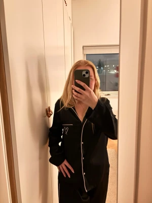Svart sidenpyjamas med ljusrosa detaljer - Snygg svart pyjamas i siden med ljusrosa kontrastsömmar och knappar. Setet har långärmad skjorta med bröstficka och byxor med raka ben. På bröstfickan finns ett litet vitt hjärta broderat. Perfekt för mysiga kvällar hemma. Nypris ca 1500kr jag är normalt strl S och passar perfekt 