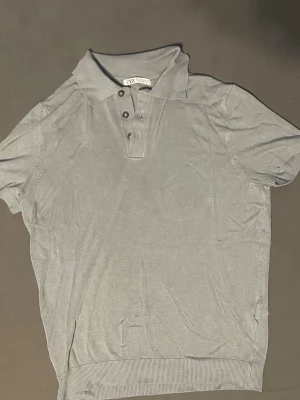 Grå pikétröja från Zara - Snygg grå pikétröja från Zara med klassisk krage och tre knappar framtill. Tröjan har korta ärmar och ribbade muddar vid ärmslut och nederkant. Perfekt för en stilren och avslappnad look.