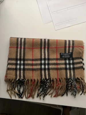 Beige rutig halsduk från Burberry - Klassisk beige halsduk från Burberry med ikoniskt rutmönster i svart, vitt och rött. Halsduken har fransar i ändarna och är tillverkad i mjukt material, perfekt för att ge din outfit en lyxig touch.
