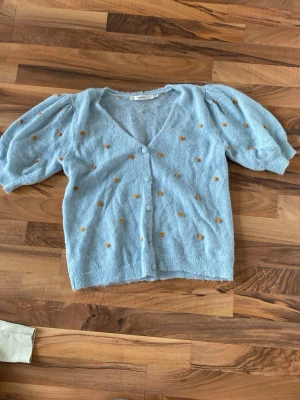 Ljusblå kofta med puffärm och prickar - Superfin ljusblå kofta från Monki med puffärmar och söta orange broderade prickar. Koftan har v-ringning, knäppning framtill och är stickad i ett mjukt material. Perfekt för att liva upp din outfit med en lekfull touch. Står ingen storlek men uppskattar den till en storlek M/L