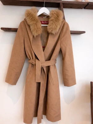 Kamelbrun kappa Max Mara - Snygg kamelbrun kappa från Max Mara med huva och bred krage i fuskpäls. Kappan har knytskärp i midjan och två fickor framtill. Lång modell som ger en klassisk och lyxig look, perfekt för kyliga dagar.