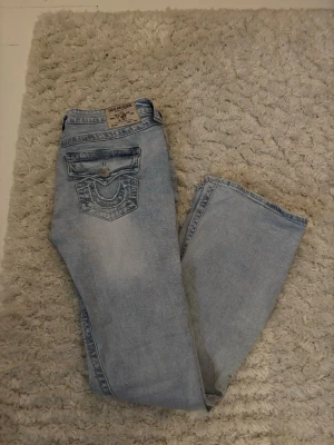 Ljusblå jeans från True Religion - Säljer min tjejs jätte fina ljusblå jeans från True Religion med klassisk broderad bakficka och silverfärgad knapp. Jeansen har en rak passform och tydliga detaljer på fickorna. Perfekta för en avslappnad och trendig stil.