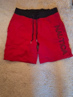 Röda shorts från Nautica med snörning - Snygga röda shorts från Nautica med svart resårmidja och dragsko. Stora bokstäver med NAUTICA tryckt längs ena benet. Perfekta för chill dagar eller träning. Mjukt material och avslappnad passform.