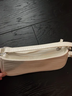 Vit axelväska i krokopräglat material - Snygg vit axelväska med krokodilpräglad yta och smal axelrem. Väskan har dragkedja upptill och en stilren, avlång form. Perfekt för dig som gillar trendiga accessoarer med lite extra edge. Pris kan diskuteras🥰