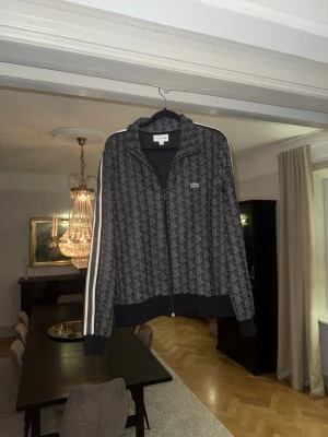 Lacoste mönstrad ziptröja - ( STORLEK L MEN PASSAR M)Snygg ziptröja från Lacoste i mörkblått med vitt mönster och vita ränder längs ärmarna. Tröjan har dragkedja framtill, ribbade muddar och klassisk Lacoste-logga på bröstet. Perfekt för en sportig och trendig look.