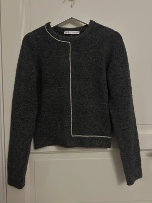 Mörkgrå stickad tröja från Zara - Snygg mörkgrå stickad tröja från Zara med vit kontrastsöm som går längs halsringningen och ner över framsidan. Tröjan har rund hals, långa ärmar och en rak passform. Perfekt för dig som gillar minimalistisk och modern stil.