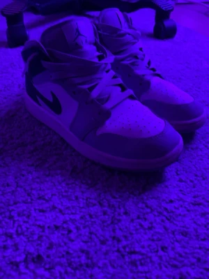 Nike Air Jordan 1 Mid sneakers - Säljer ett par Nike Air Jordan 1 Mid sneakers med klassisk siluett. Skorna har vit bas med gråa och svarta detaljer samt den ikoniska swoosh-loggan på sidan. Ovandelen är i läder och de har snörning framtill. Perfekta för dig som gillar streetwear och sportig stil.