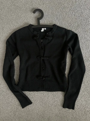 Svart kofta med knyt från Nelly - En svart kofta/tröja ifrån Nelly med fina rosetter och knallar framtill. Den går att knäppa upp helt och ha som kofta också! ❣️