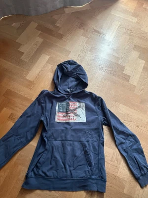 Mörkblå hoodie med USA-flagga - Snygg mörkblå hoodie från Replay & Sons med stor ficka framtill och tryck av en amerikansk flagga på bröstet. Hoodien har huva och är tillverkad i mjukt bomullsmaterial. Perfekt för en avslappnad streetwear-look.