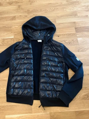 Svart cardigan med huva från Moncler - Snygg svart cardigan från Moncler med quiltad front och rygg i glansigt material, samt mjuka ärmar och huva. Jackan har dragkedja framtill, fickor och en Moncler-logga på ärmen (med en liten ficka). Perfekt för dig som vill ha en trendig look.