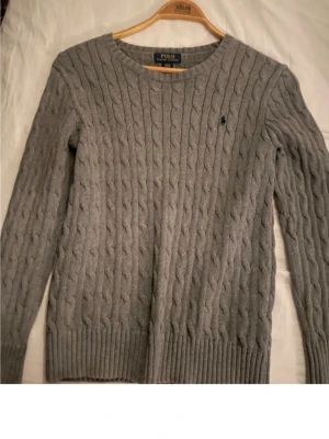 Grå kabelstickad tröja Polo Ralph Lauren - Kabelstickad grå tröja från Polo Ralph Lauren med rund halsringning och diskret broderad logga på bröstet. Långa ärmar och ribbade muddar vid ärmslut och nederkant. Perfekt för lager-på-lager och klassisk stil.