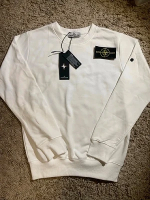 Vit sweatshirt från Stone Island - Vit sweatshirt från Stone Island med klassisk rund halsringning och ribbade muddar. Tröjan har en avtagbar logotyp-patch på ärmen och är tillverkad i mjukt material som känns skönt mot huden. Perfekt för en clean och stilren look.