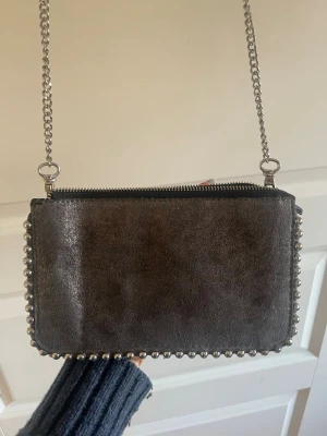 Mörkgrå axelväska med kedjerem - Snygg mörkgrå axelväska i mockaliknande material med silverfärgade nitar längs kanterna och en lång kedjerem. Väskan har dragkedja upptill och en stilren, rektangulär form som ger en cool vibe till din outfit.