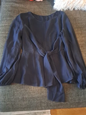 Mörkblå topp med knyt i midjan - Snygg mörkblå topp med långa ärmar och knytband i midjan. Modellen har rund halsringning och är tillverkad i ett mjukt tyg som ger en bekväm passform. Perfekt för att skapa en stilren look.