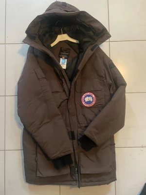  Canada Goose jacka - Säljer en brun parkas från Canada Goose med ikonisk röd och vit logga på bröstet. Jackan har huva, flera stora fickor och muddar vid ärmsluten. Perfekt för kalla dagar och riktigt varm tack vare det tjocka fodret.