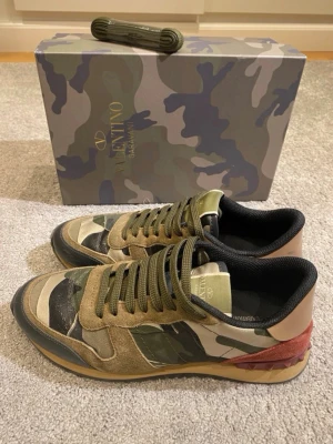 Valentino camo sneakers med mockadetaljer - Snygga sneakers från Valentino med camouflagemönster i grönt, beige och svart. Skorna har detaljer i mocka och läder samt en unik röd detalj vid hälen. Klassisk låg modell med gröna skosnören och svart innersula. Perfekta för dig som vill sticka ut med stil.