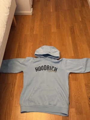 Ljusblå hoodie från Hoodrich - Snygg ljusblå hoodie från Hoodrich med svart broderad logga på bröstet och en stor ficka framtill. Hoodien har en klassisk huva och är tillverkad i mjukt material som passar perfekt för en avslappnad stil. Xl i barn
