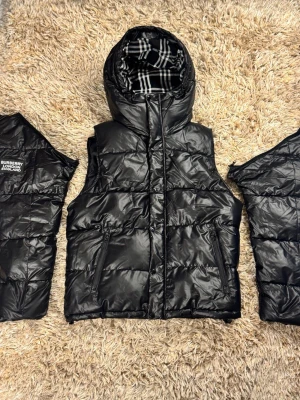 Svart dunväst från Burberry - Snygg svart dunväst från Burberry med glansig finish och klassiskt rutigt foder i huvan. Västen har två dragkedjefickor framtill och hög krage. Perfekt för lager-på-lager och streetstyle.