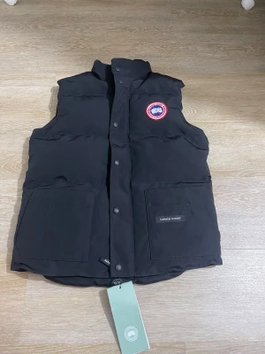 Svart dunväst från Canada Goose - Snygg svart dunväst från Canada Goose med klassisk logga på bröstet och två stora fickor framtill. Västen har hög krage och knäppning med tryckknappar. Perfekt för lager-på-lager och håller dig varm under kyliga dagar.