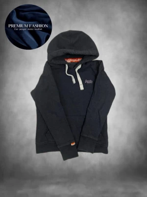 Mörkblå hoodie från Superdry - Mörkblå hoodie från Superdry med vit dragsko i huvan och liten logga broderad på bröstet. Klassisk känguruficka framtill och ribbade muddar vid ärmslut och nederkant. Perfekt för en avslappnad och stilren look.